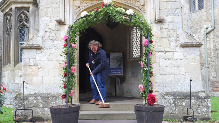 Volunteer sweeping the ground in front of the house ready for the house opening at Anglesey Abbey. The lady is sweeping under an arch of dahlias.